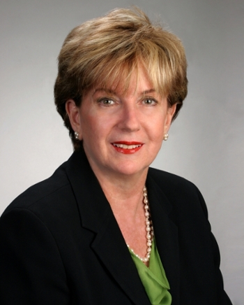 Portrait of a woman in a black suit and green blouse wearing a pearl necklace on a gray background